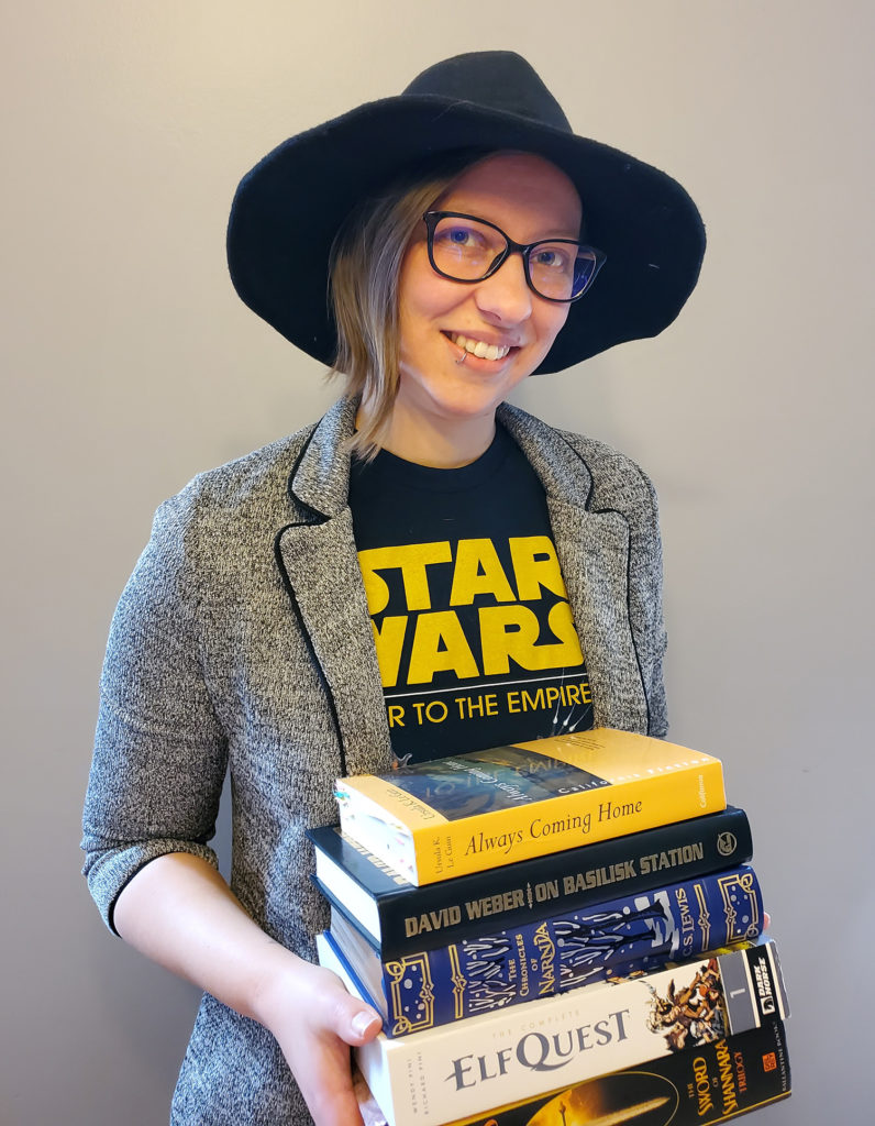 A photograph of Kelly the Book Witch holding a stack of their favorite books and wearing a black witch hat.