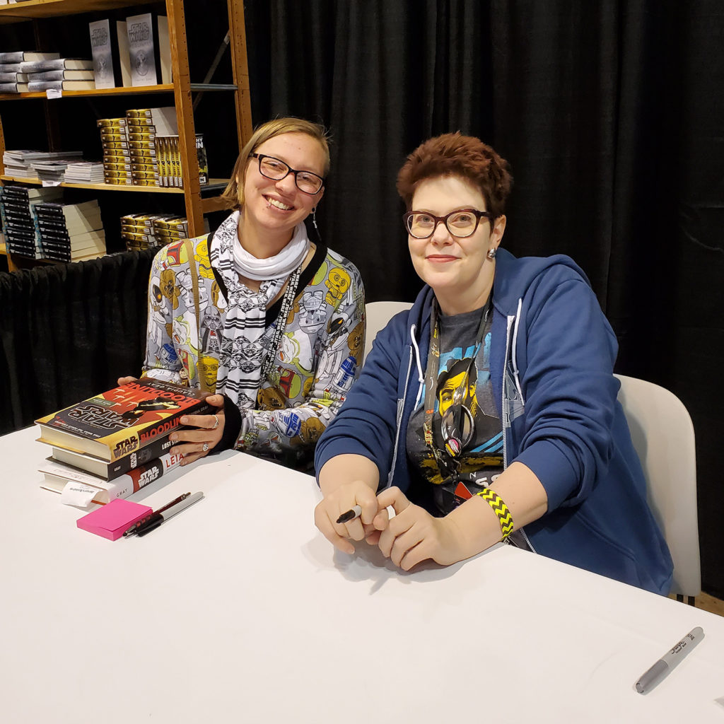 Kelly sitting next to Claudia Gray at a book signing, holding a stack of Claudia's Star Wars books.