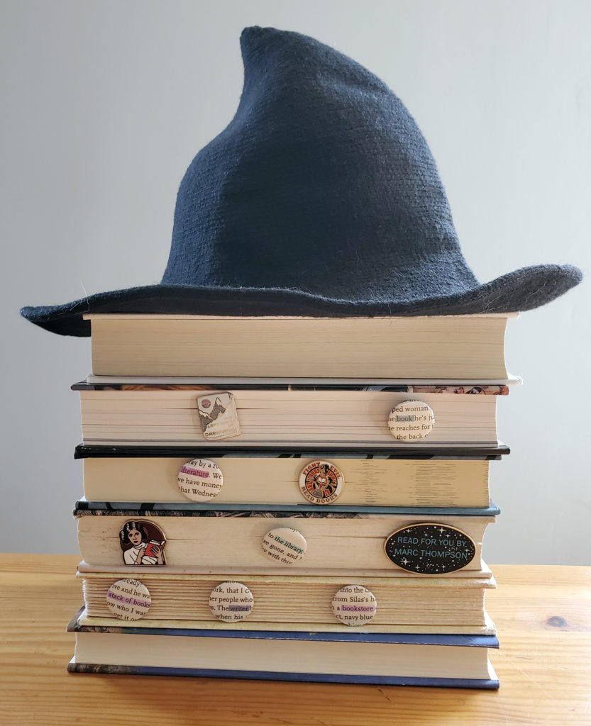 A stack of books, pages face out, with a witch hat on top of the stack. Pins and buttons are held between the pages.