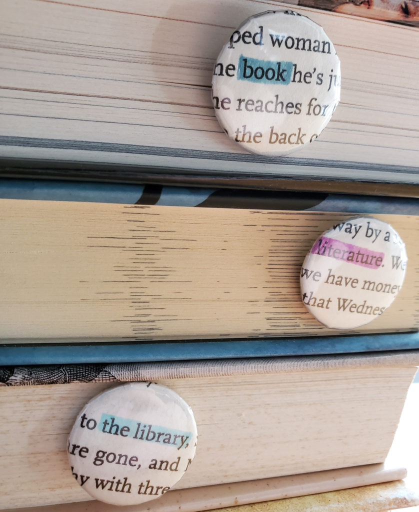 Three pinback buttons made from recycled book pages with highlighted words: book, literature, and the library. The pins are photographed against a background of stacked books.
