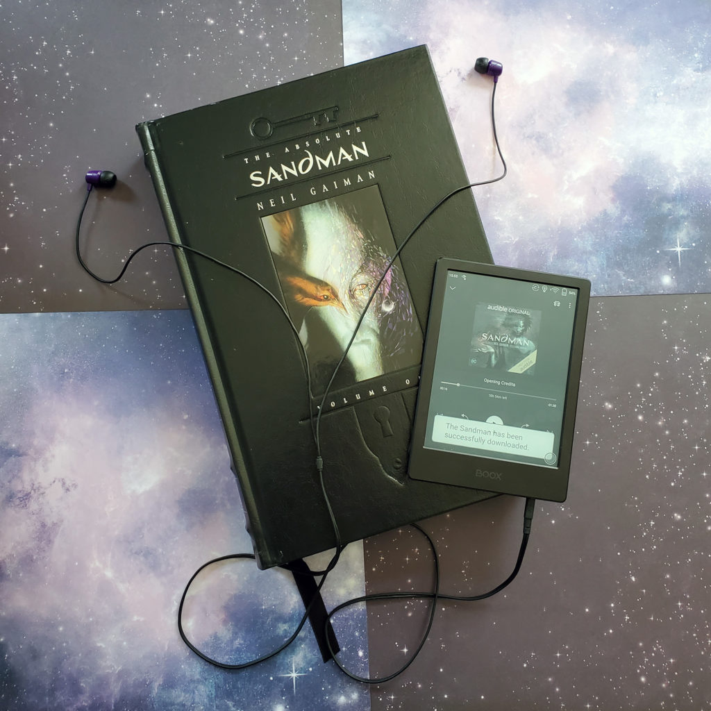 The Absolute Sandman volume 1 on a starry background, with an eReader playing the Sandman audio book on top of it.