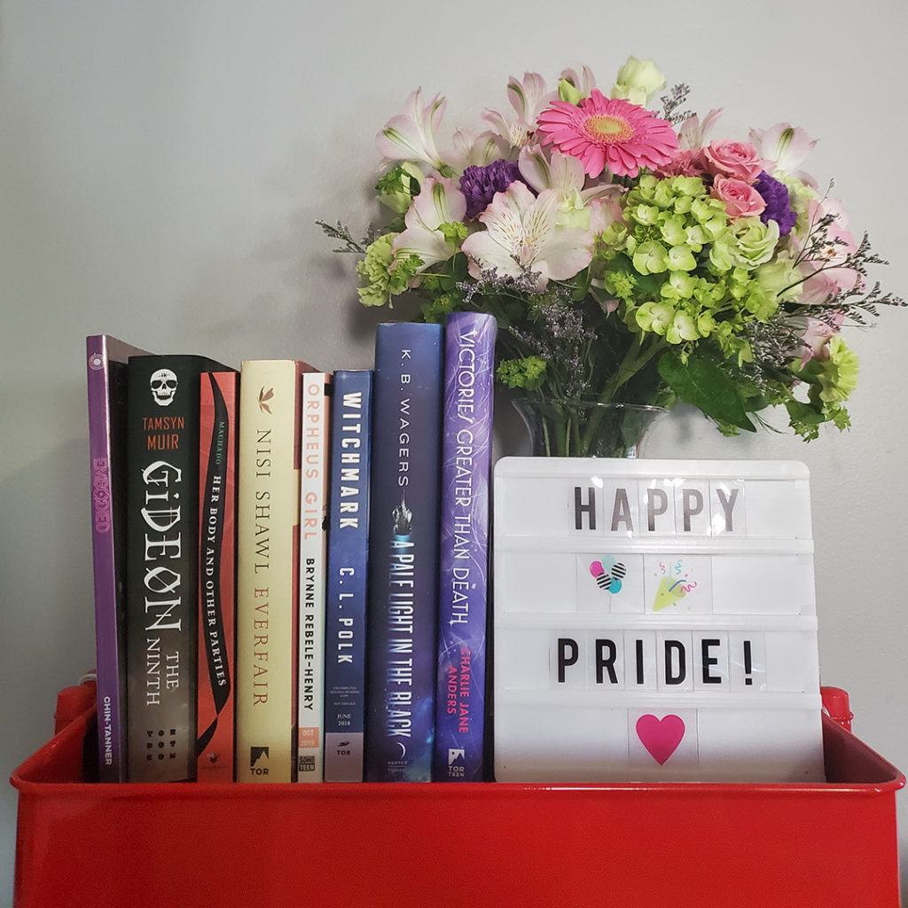 8 books next to a light box that reads "Happy Pride" with a bouquet of flowers in the background.