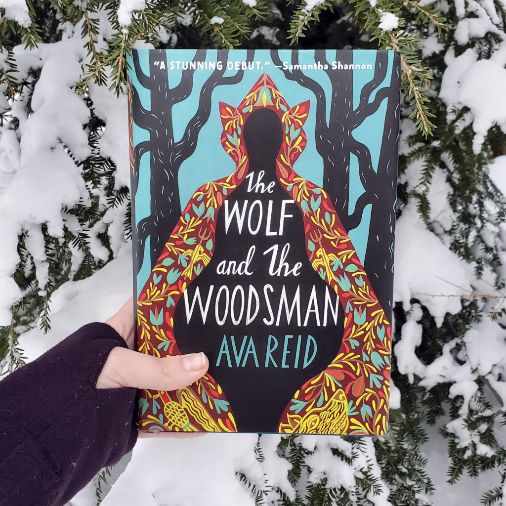 Photograph of me holding Ava Reid's The Wolf and the Woodsman in front of a snow covered Eastern Hemlock tree. 