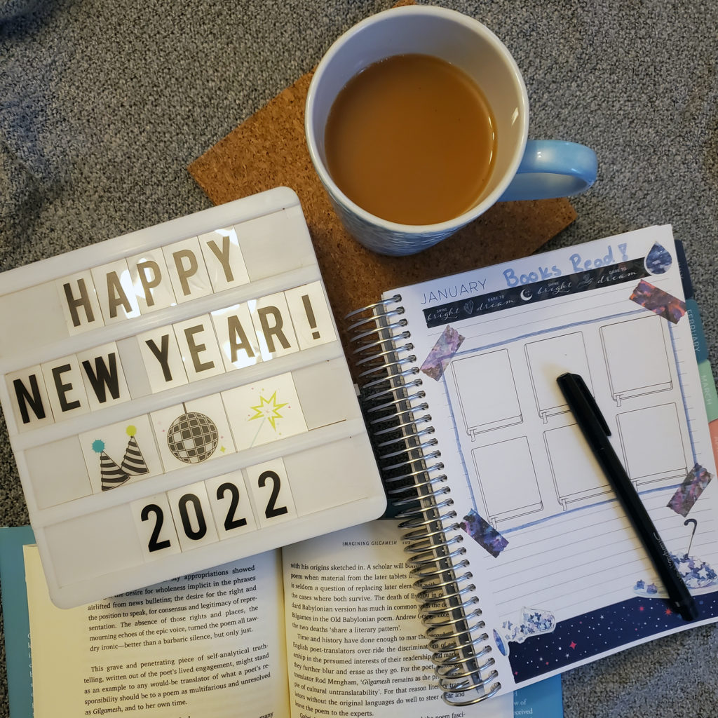 A photo of a planner, an open book, and a board that says "Happy New Year 2022" next to a mug of tea, all one a gray knitted fabric background.