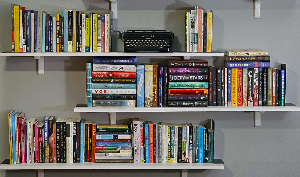 A photo of three shelves full of books. A typewriter sits on the middle in the top shelf, with books on either side. One reading goal alternate can be to read books from your TBR pile